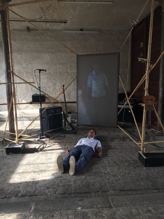 Dominik lying on a stone floor surrounded by bamboo structure, in front of a video screen showing him lying on a stone floor.