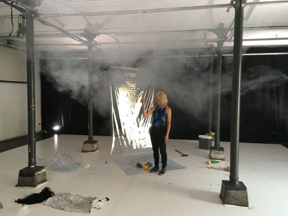 Elina in blonde wig in a theatre space, white floor, scattered elements, a transparent plastic foil and faint smoke in the air.